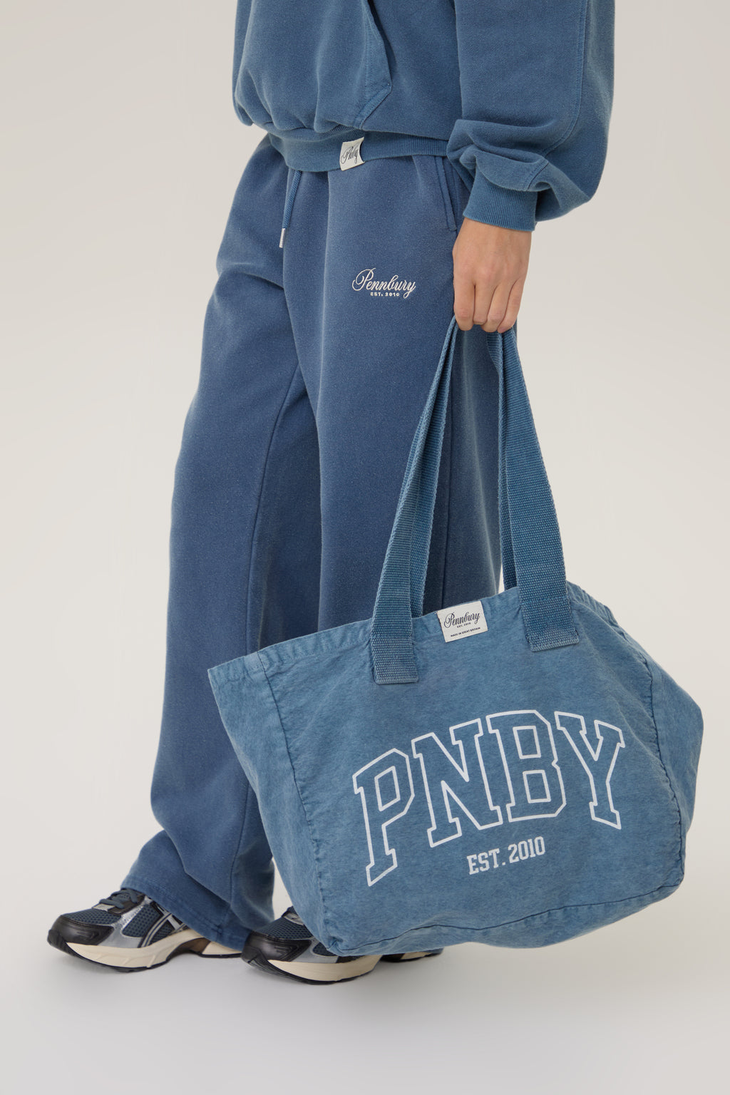 Person wearing blue pants and holding a blue bag with 'PNBY' branding on a light gray background