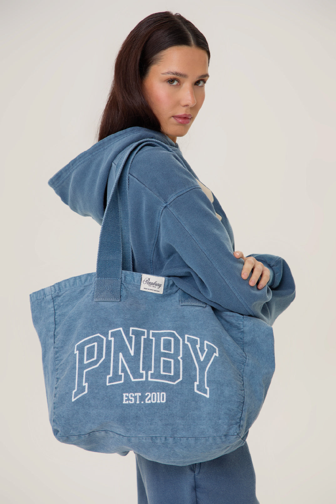Woman wearing a blue hoodie with a tote bag featuring 'PNBY' branding on a plain background