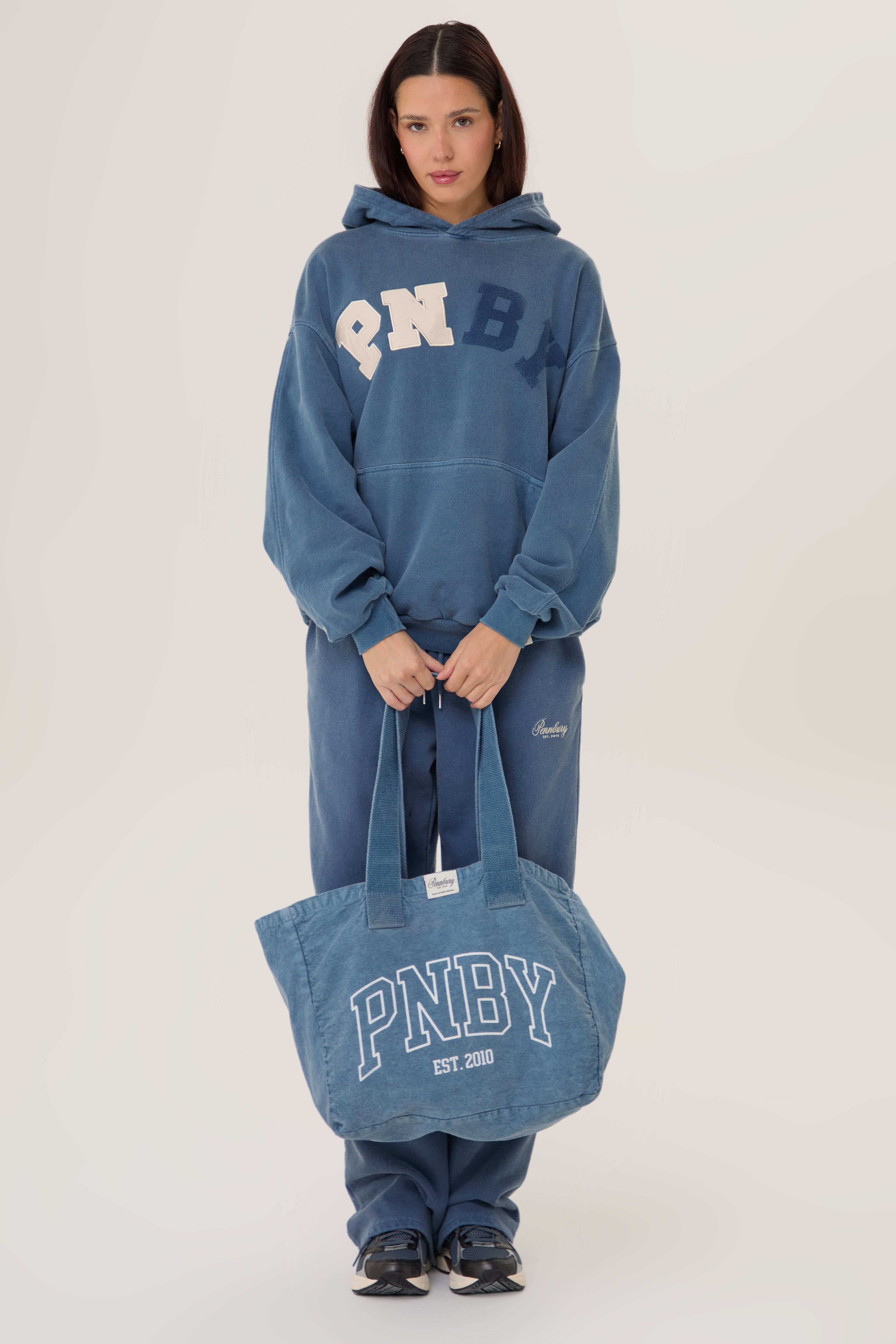 Person wearing a blue hoodie and pants holding a matching tote bag with 'PNBY' branding on a white background.